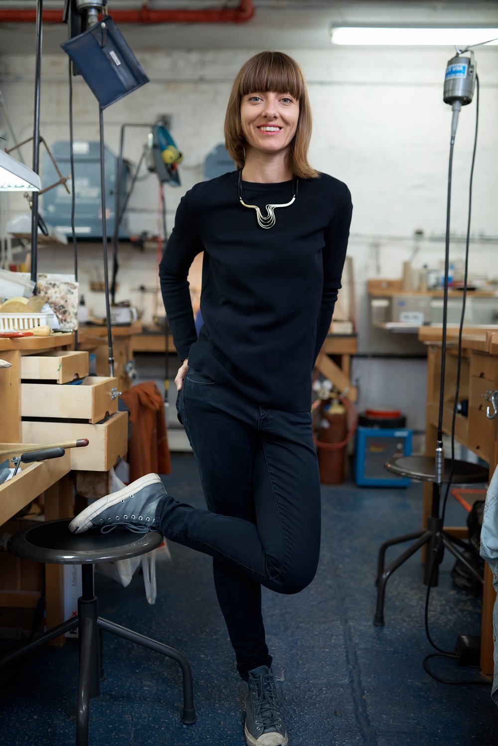 Full-length environmental portrait of a jewelry designer standing at her workbench in a Brooklyn studio