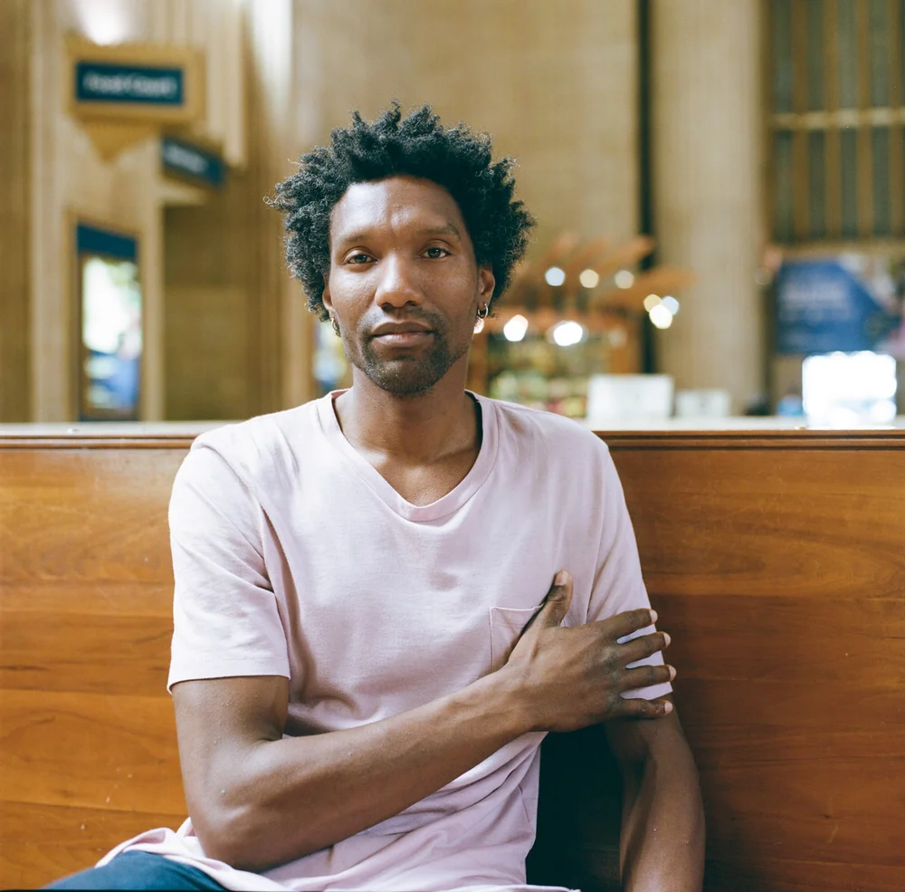 Editorial portrait of a man seated on a wooden bench inside a railway concourse