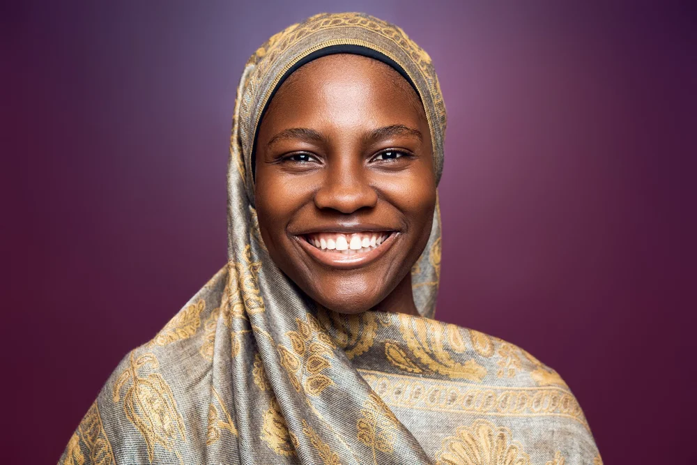 Smiling studio headshot of a woman in a gold patterned headscarf against a purple background