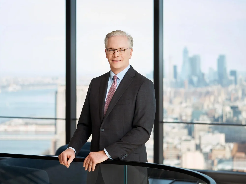 Executive portrait of a managing partner in a dark suit and glasses, photographed in a Manhattan office with a city view