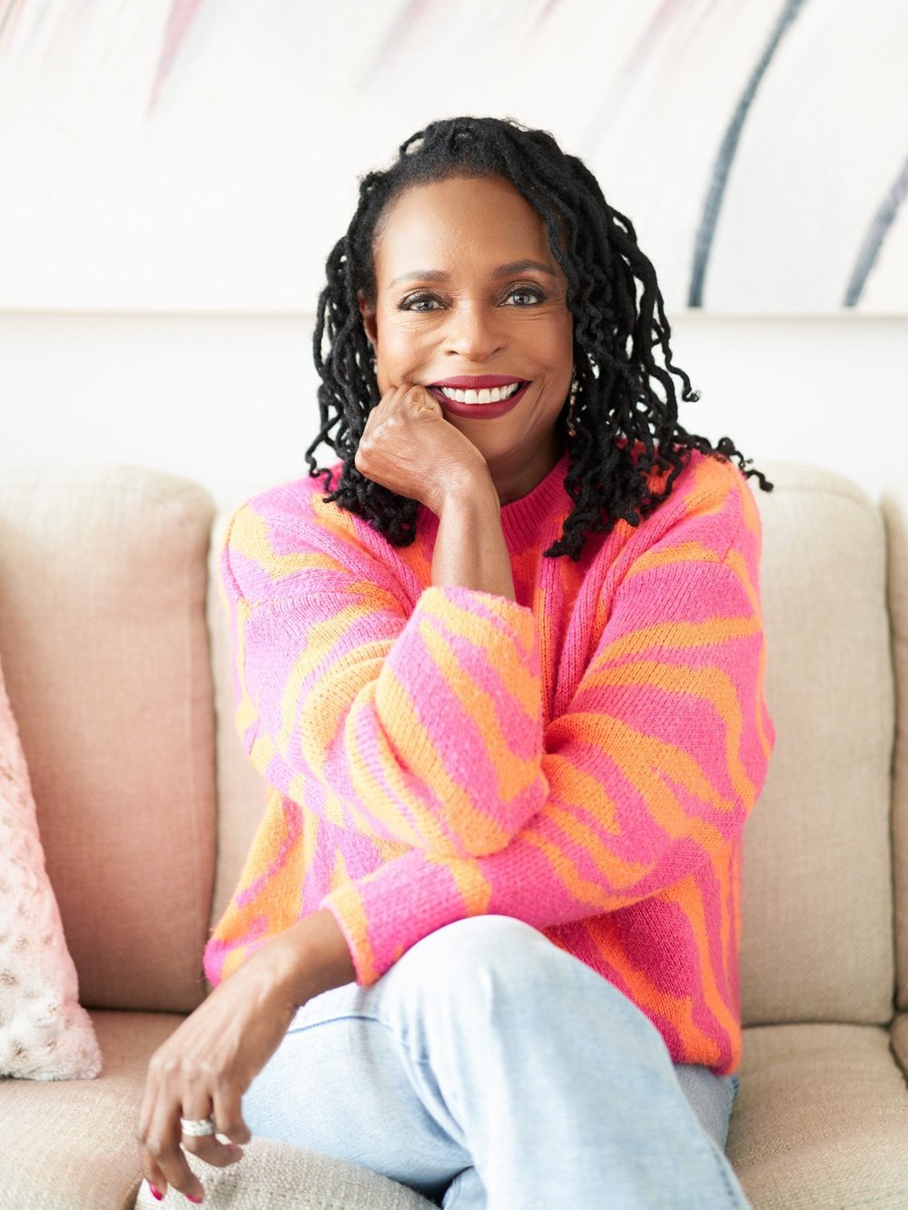Personal branding portrait of a smiling woman in a bright pink and orange sweater seated on a sofa