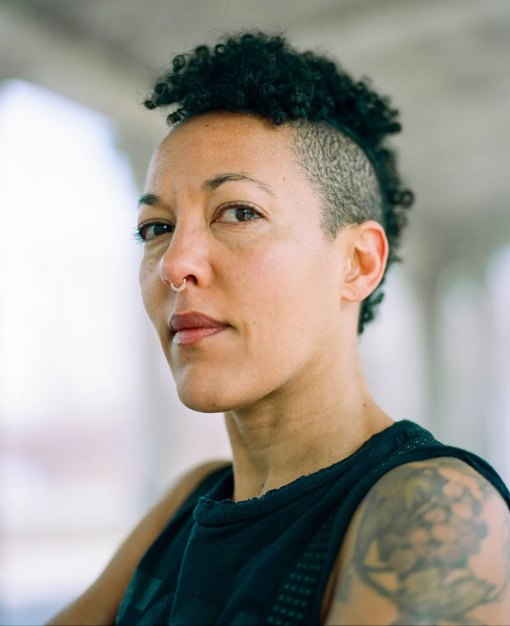 Natural-light film portrait of a person with short curly hair, a nose ring and a visible shoulder tattoo