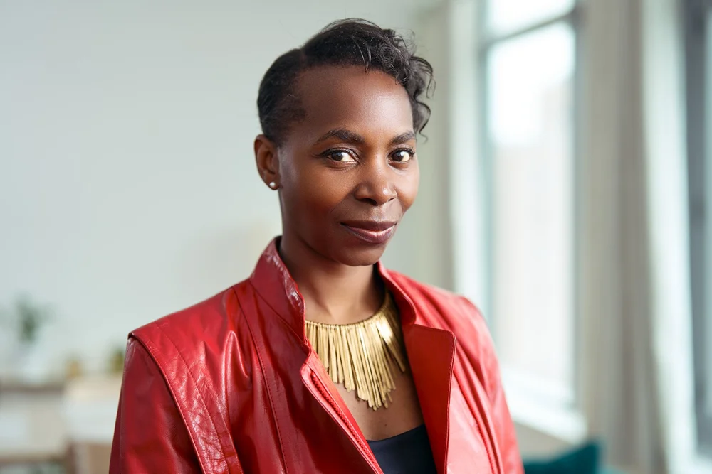 Editorial portrait of a woman in a red leather jacket, photographed beside a bright apartment window