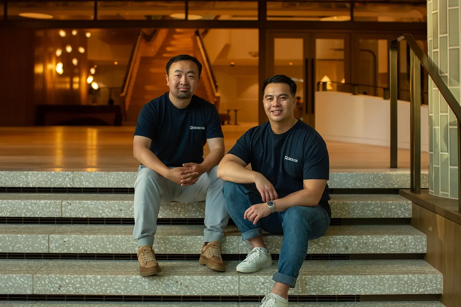 Abacor founders seated on exterior steps wearing branded company t-shirts