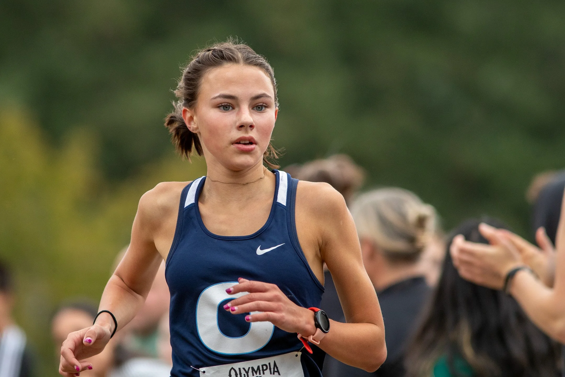 Girl in navy Olympia jersey with white O logo running past spectators at the 2026 Olympia XC Classic.