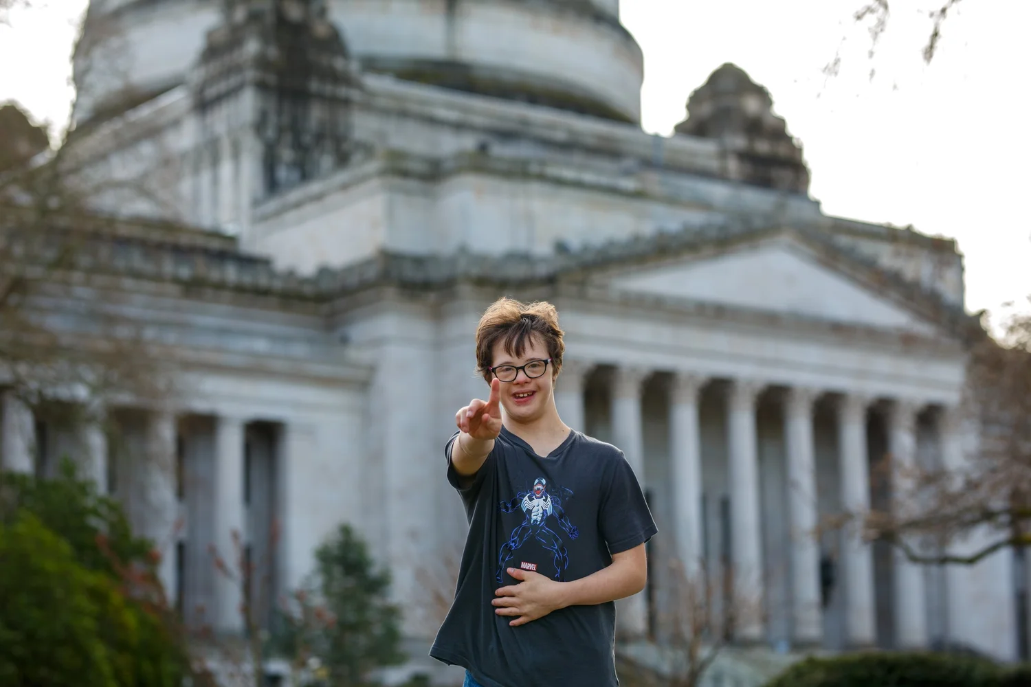 Person in a navy shirt pointing at the camera in front of the Legislative Building on the Washington State Capitol campus in Olympia