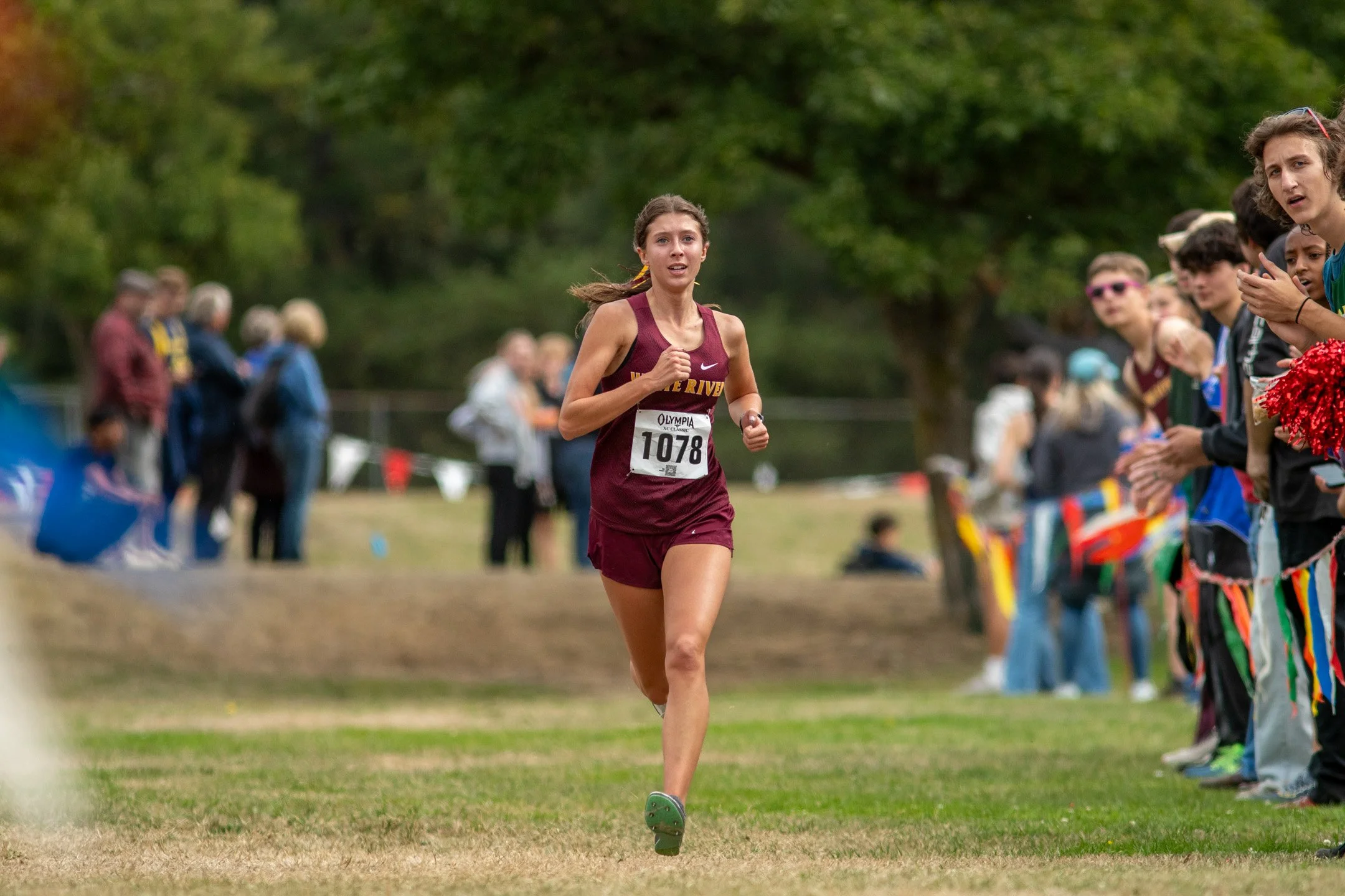Girl in maroon White River jersey, bib 1078, running past cheering spectators at the 2026 Olympia XC Classic.