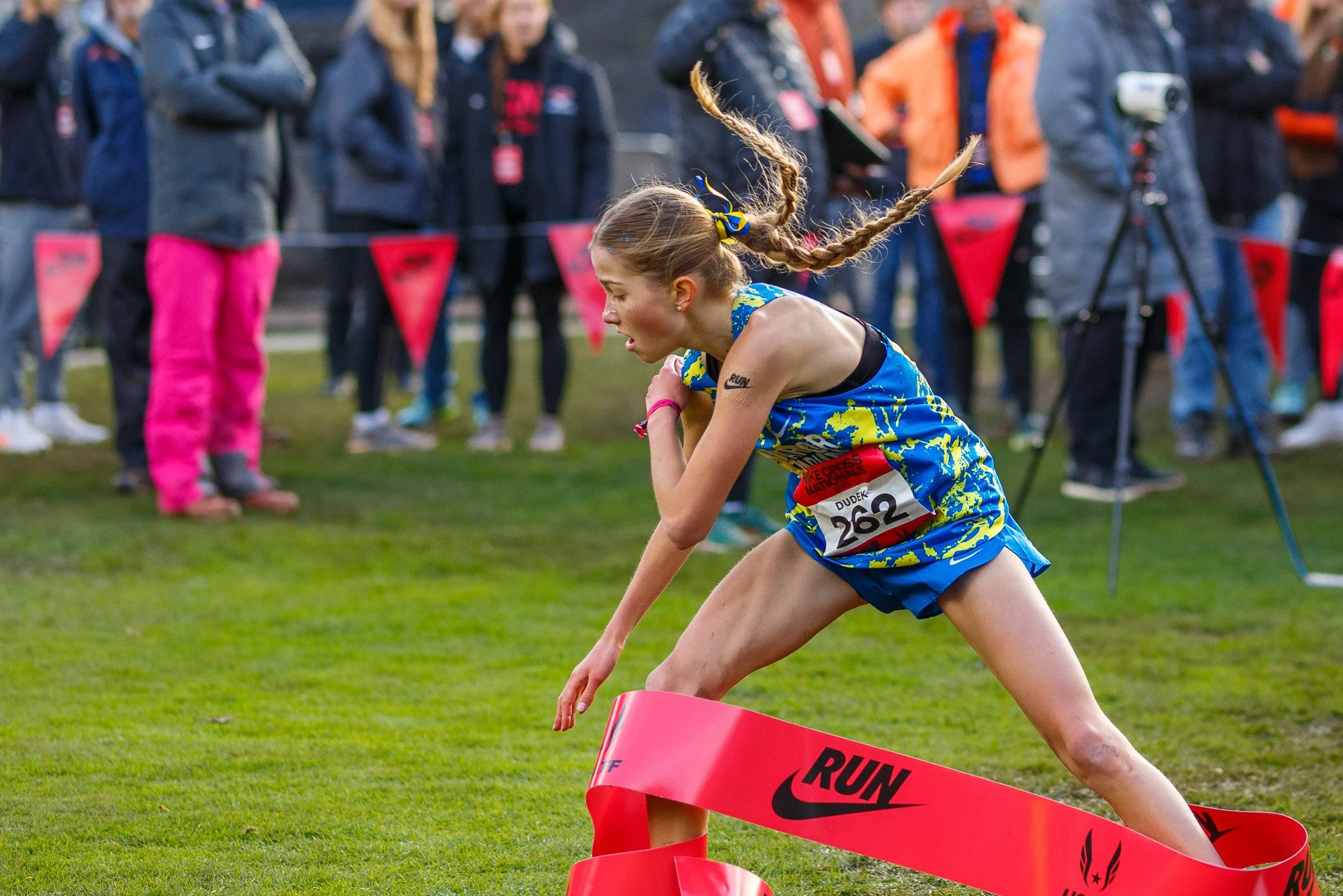 A female high school cross country runner with a braided ponytail wearing a blue and yellow uniform breaks the red Nike finish line tape. She has mud on her legs and intense focus at the 2025 Nike Cross Nationals in Portland, Oregon.