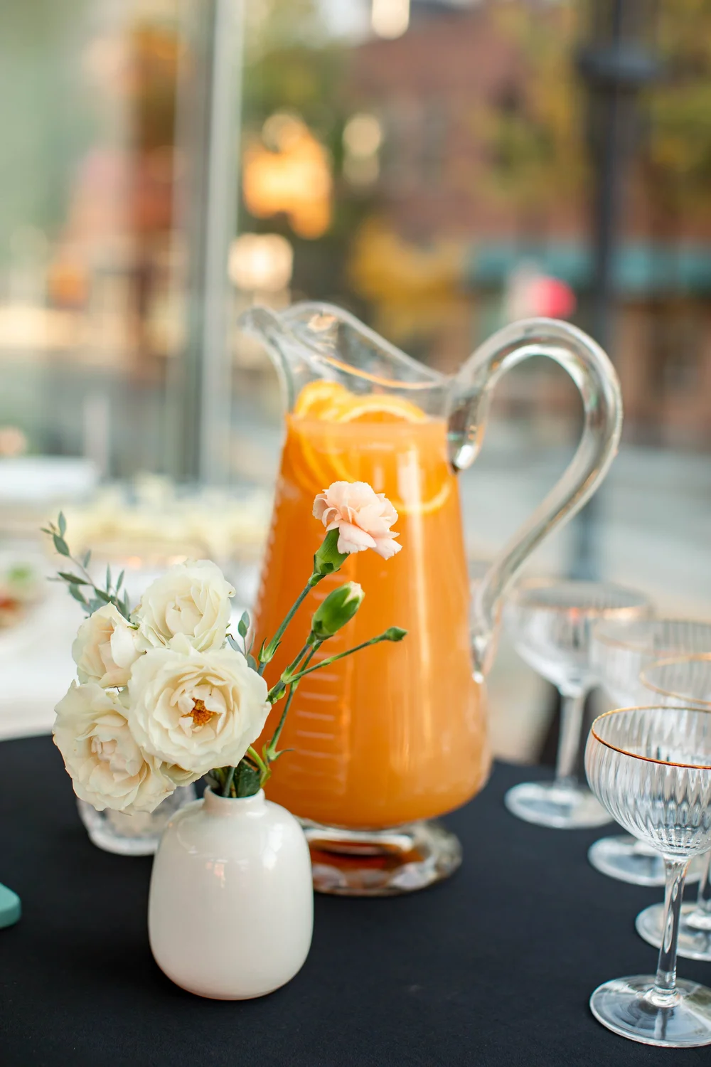 A glass pitcher with citrus beside flowers at the drinks table.