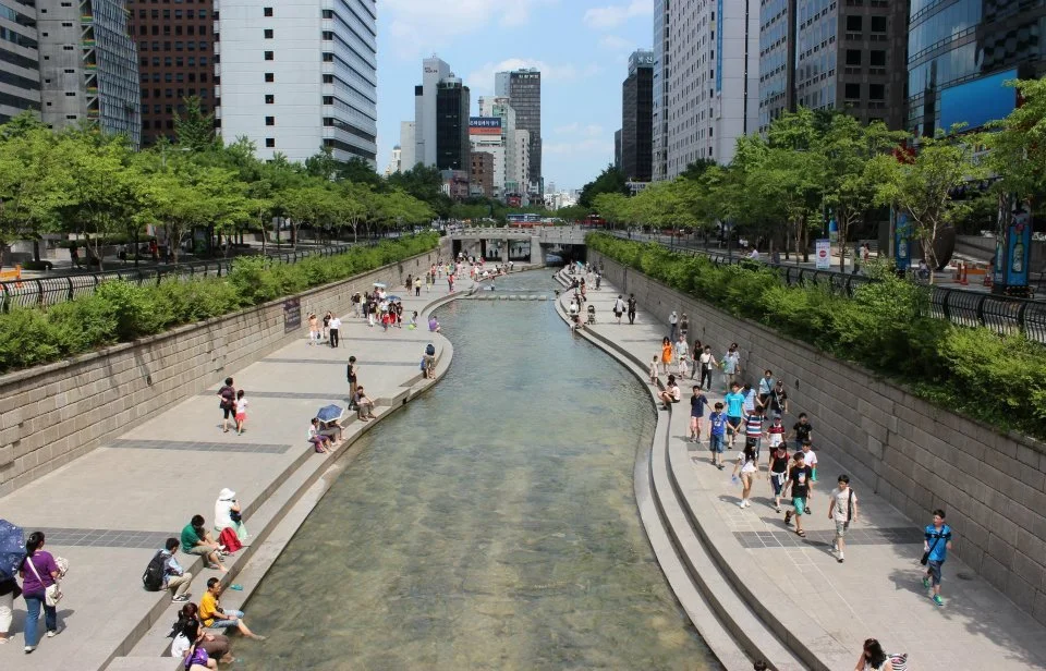 A river is surrounded by concrete steps leading down to it. People walk along the paths beside the steps while others dip their feet in the water.