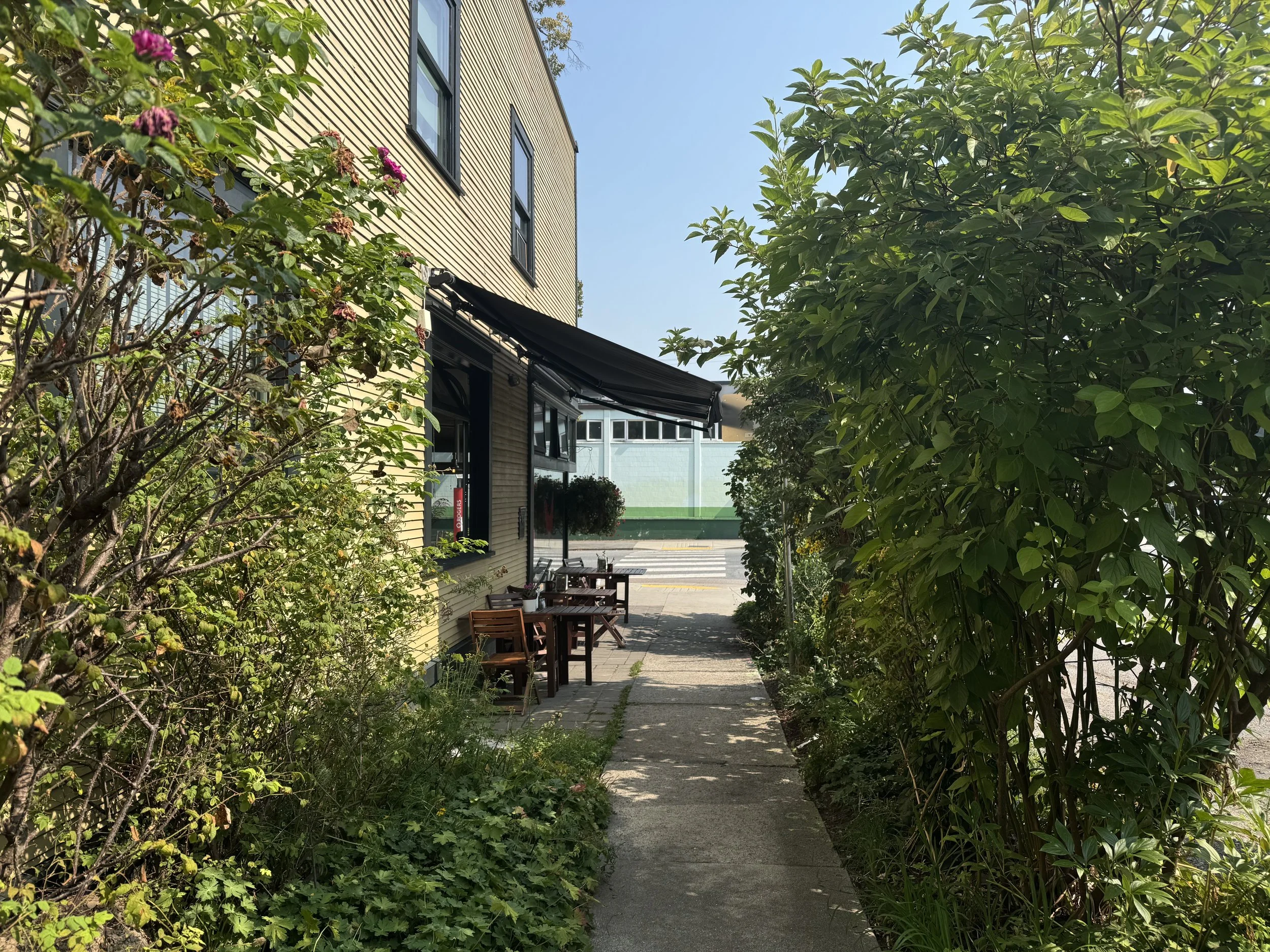 A sidewalk with gardens on both sides. At the end of a sidewalk is a yellow cafe with a black awning and outdoor seating.
