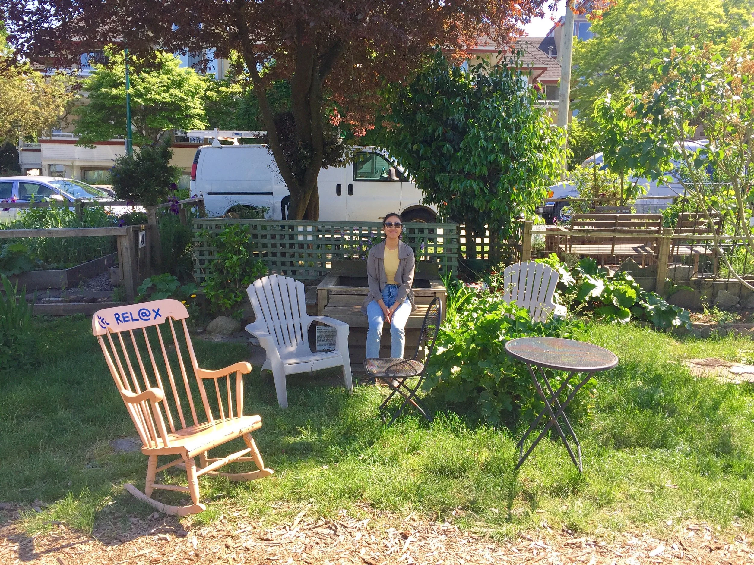 A couple of mismatched chairs in an outside greenspace. A woman sits on a chair in the centre.