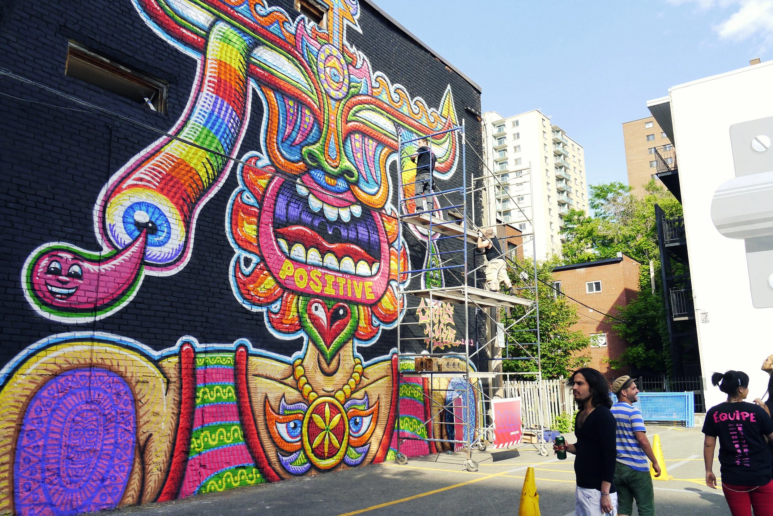 An artist stands on scaffolding and paints a colourful mural on a building. People watch from below.