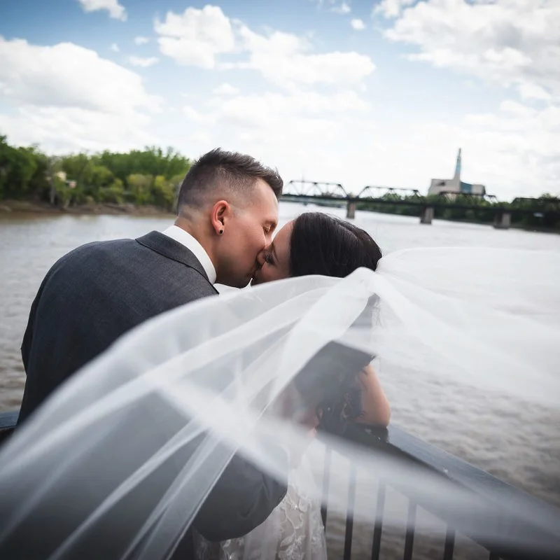 Bride and groom share a kiss on a riverside boardwalk, her veil sweeping across the frame with a railway bridge over the water behind them.