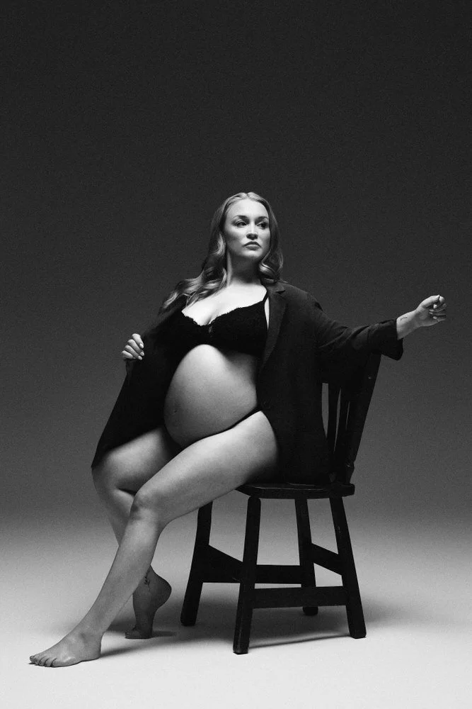 Black and white portrait of an expectant mother seated in a chair, black blazer, hands at her waist