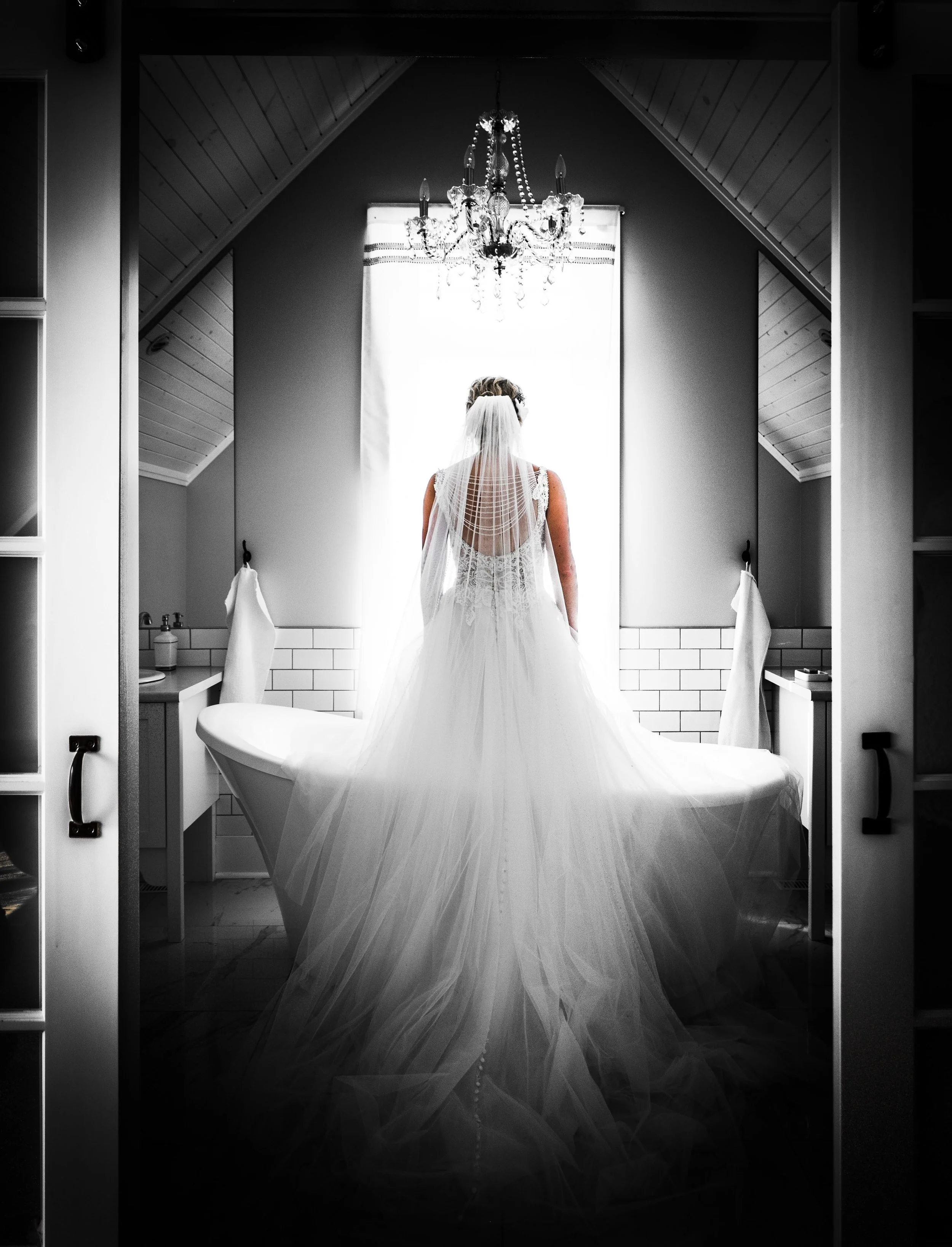 Bride photographed from behind in black and white, her cathedral veil trailing across a clawfoot tub as she looks toward the window before her Winnipeg wedding.