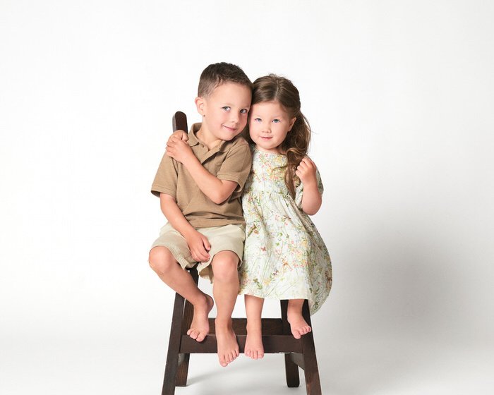 Studio sibling portrait — brother and sister seated on a wooden stool in tan and floral outfits
