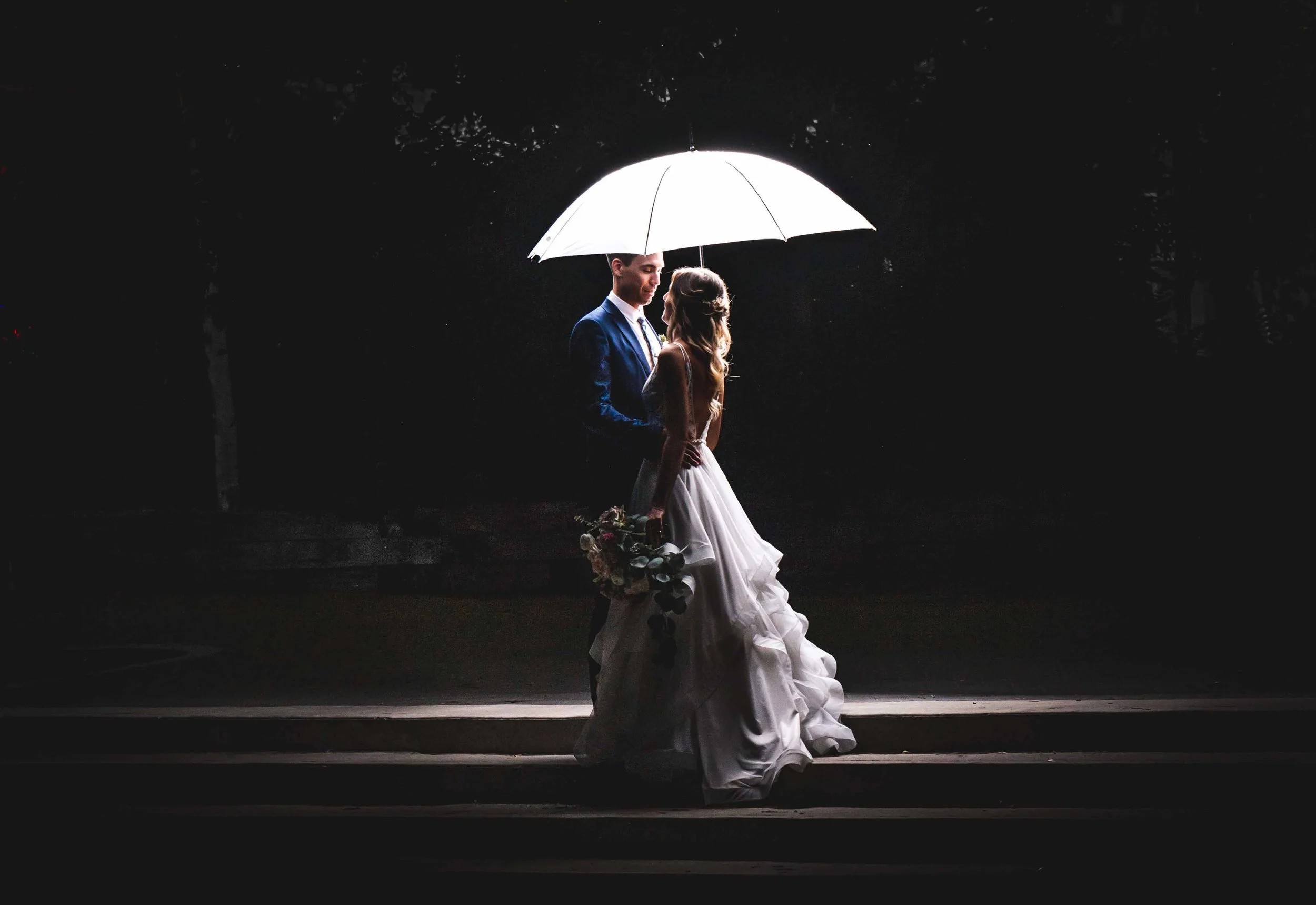 Bride and groom sharing a quiet moment under a glowing white umbrella at night, her bouquet visible against the dark surroundings.