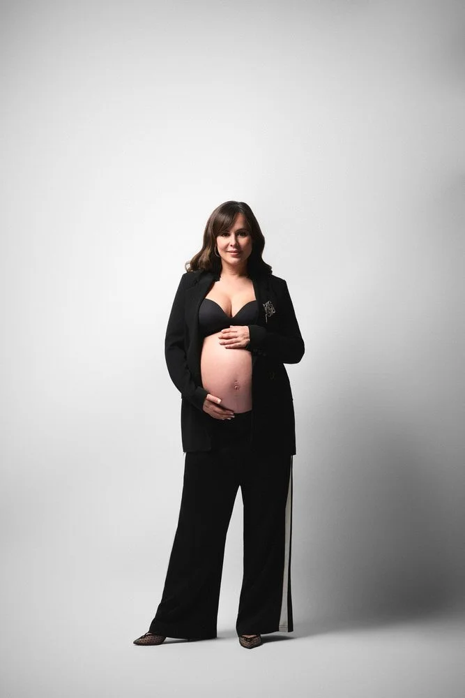 Expectant mother in a tailored black suit, blazer open, cradling her belly in a studio maternity portrait against a soft white backdrop.