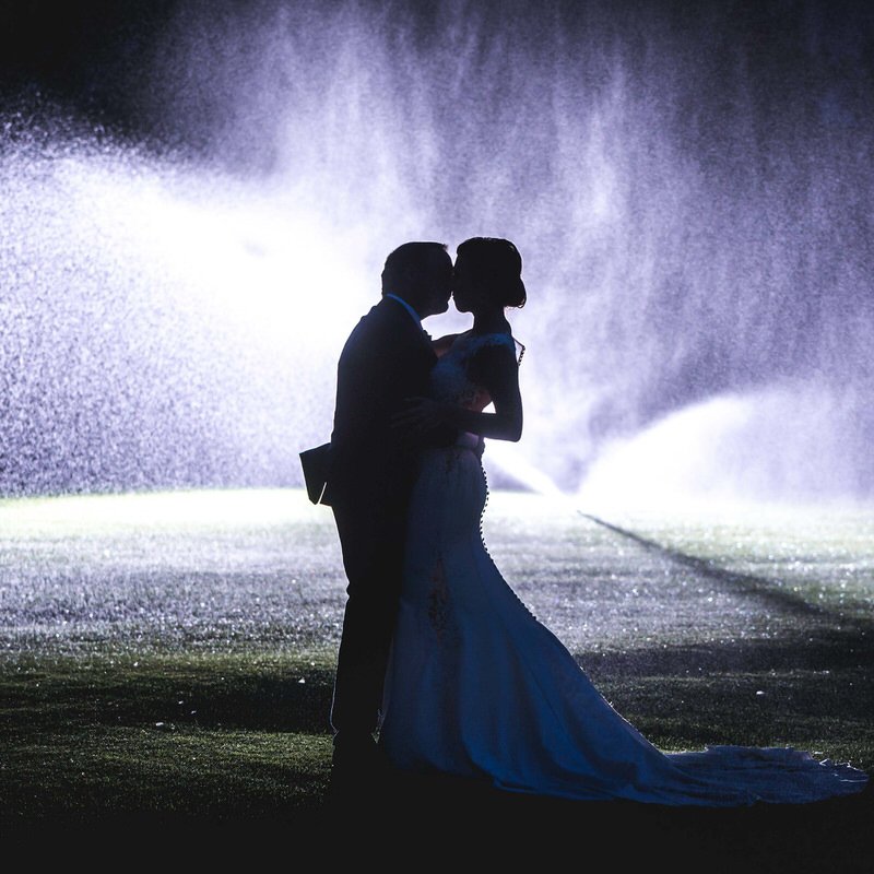 Outdoor wedding portrait — couple kissing dramatically backlit with sprinklers