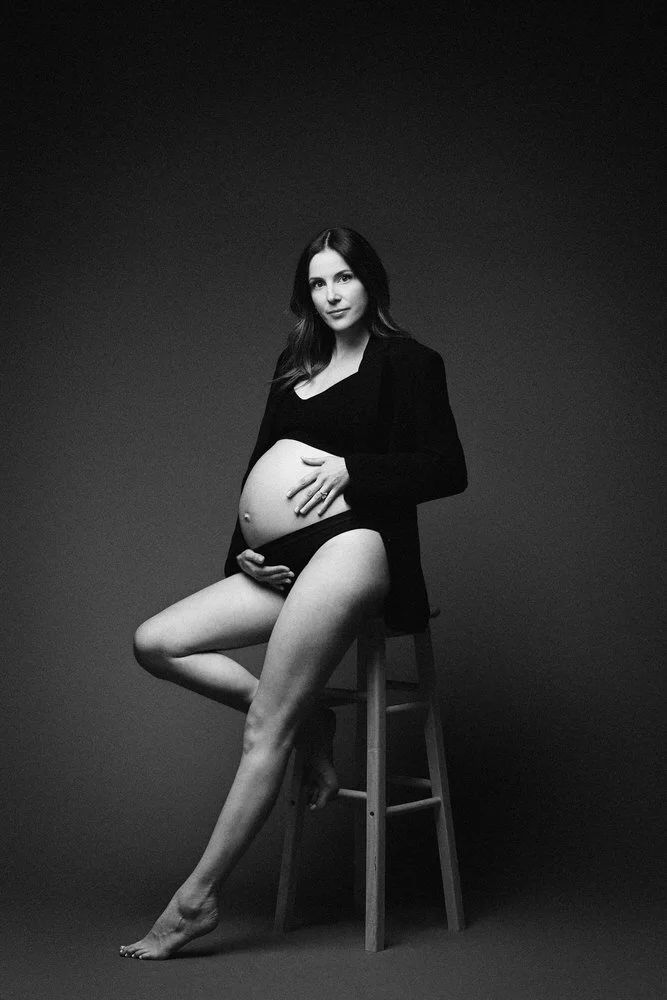 Black and white studio portrait of an expectant mother seated on a stool in a black blazer