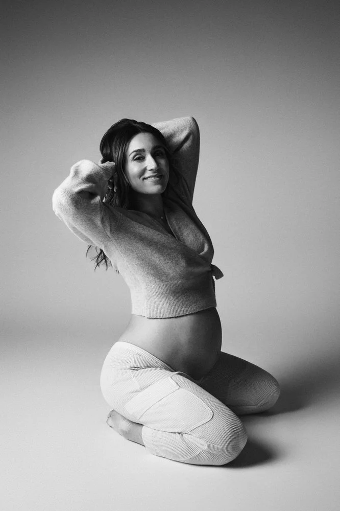 Expectant mother kneeling in an open wrap sweater, smiling, in black and white