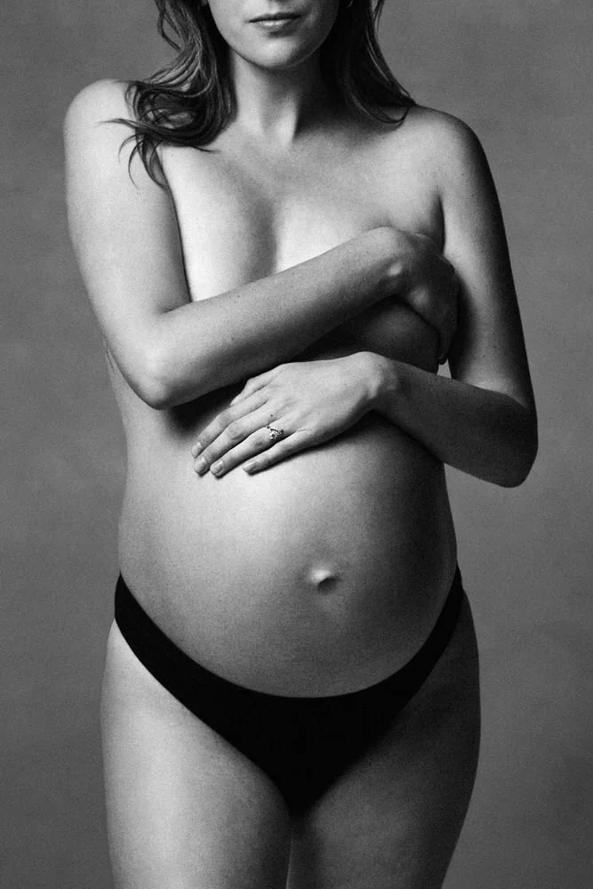 Black-and-white maternity portrait, a mother-to-be cradling her belly with crossed arms, her engagement ring catching the light.
