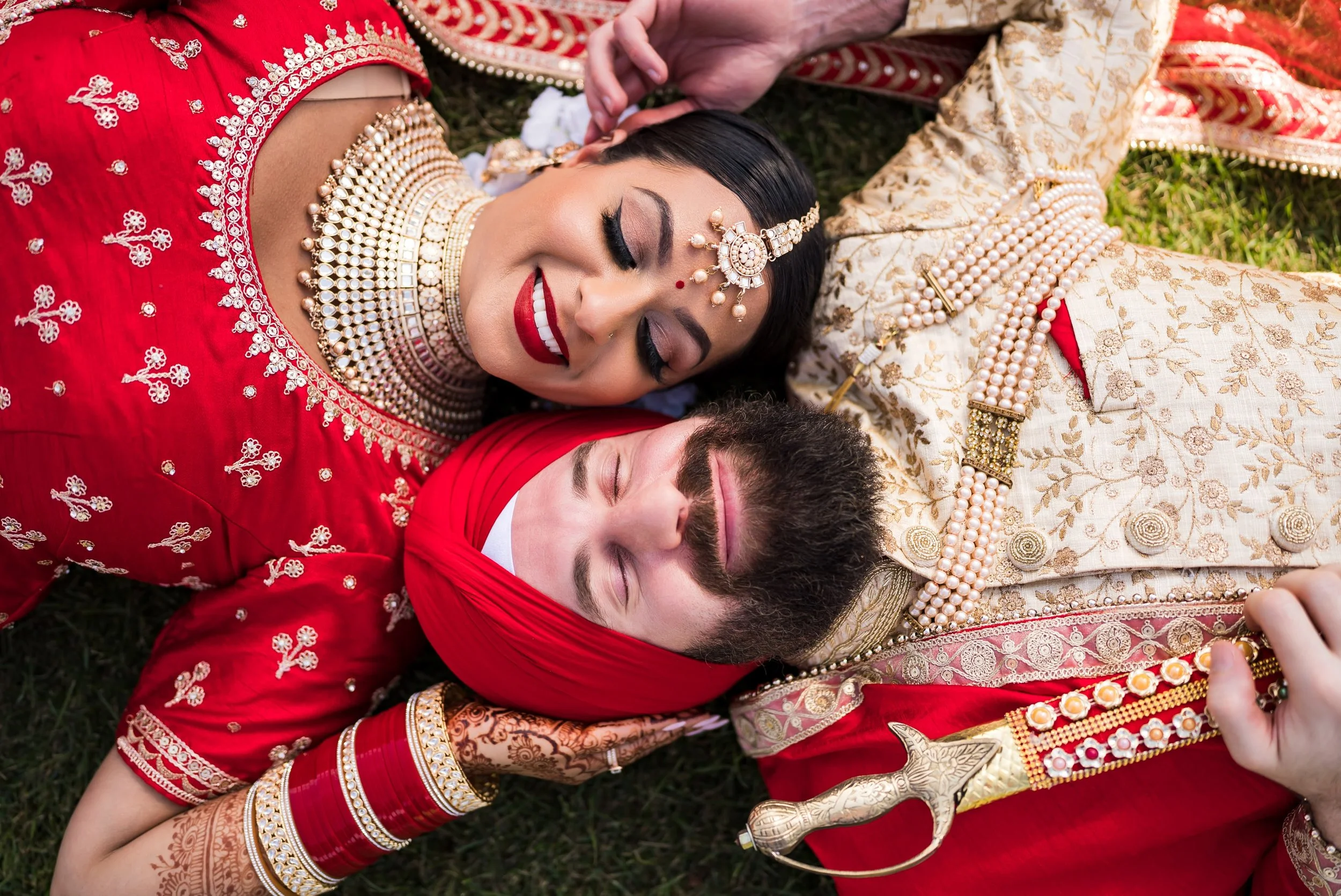 Bride and groom in traditional red and gold wedding attire lying together on the grass and laughing, jewelry and henna detailing visible.