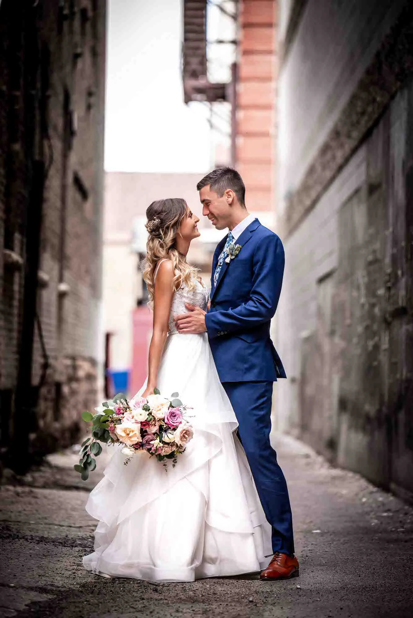 Outdoor wedding portrait — couple smiling in an alley, bride holding a bouquet, groom in a blue suit