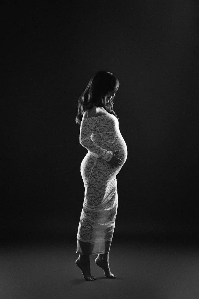 Black and white maternity portrait in a white lace dress against a dark studio backdrop
