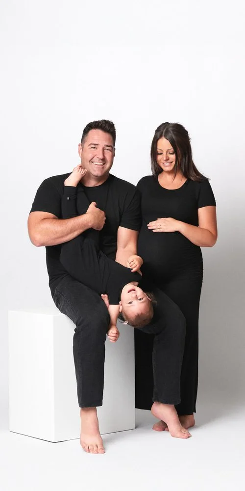 Studio family portrait — expecting couple in all black with playful toddler hanging upside down