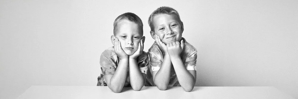 Black-and-white portrait of two brothers resting their chins on their hands at a table, one smiling and one looking on solemnly.