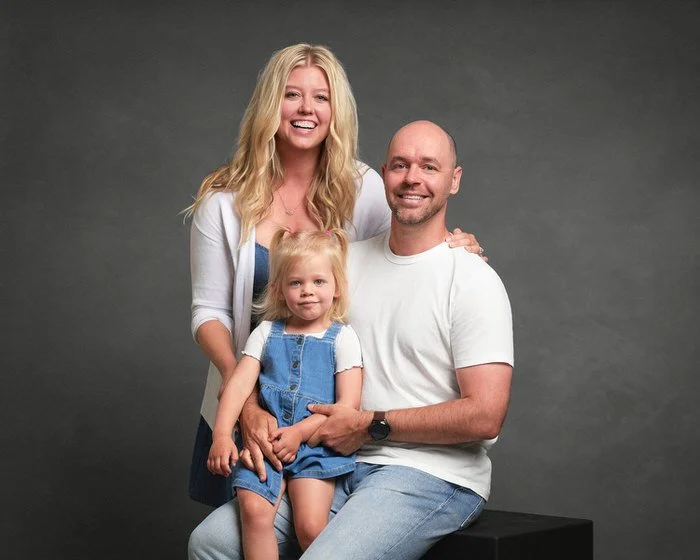 Studio family portrait — family of three in denim and white with their daughter in overalls against a dark backdrop