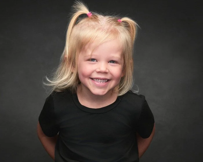 Studio portrait of a young girl with pigtails laughing in a black t-shirt, photographed against a dark backdrop.