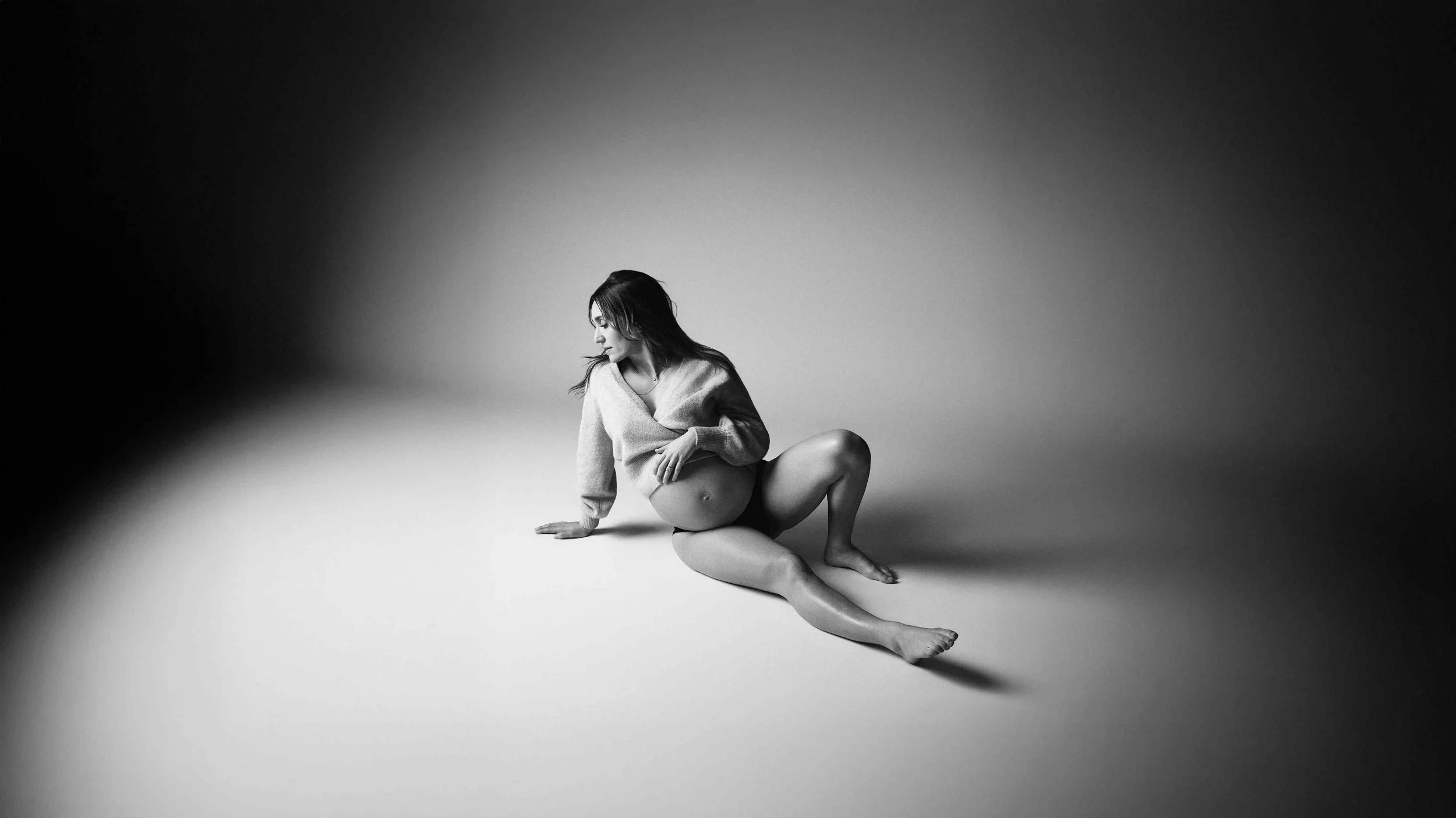 Black-and-white maternity portrait of an expectant mother reclining on the studio floor in a soft knit wrap, looking back over her shoulder.