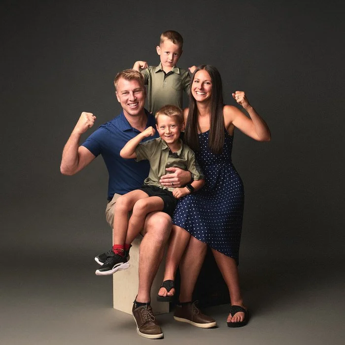 Studio family portrait — family of four in blue and olive flexing together against a dark backdrop