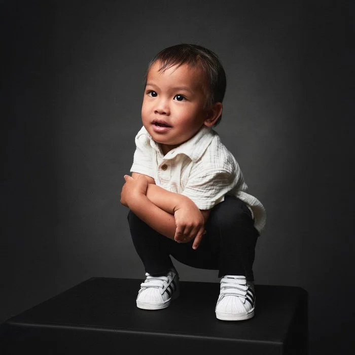 Toddler boy in a cream shirt crouched on a black studio block, arms crossed, mid-expression during a family photography session.