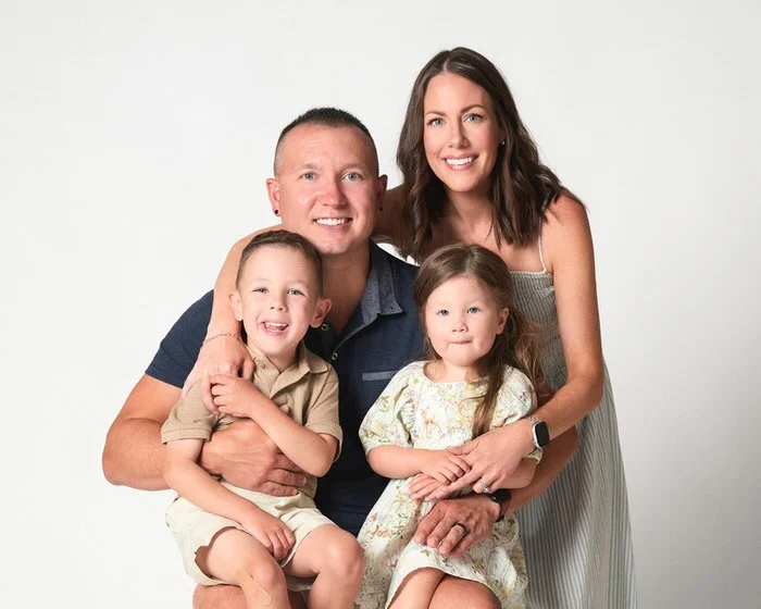 Studio family portrait of two parents and their two children, dressed in navy and sage tones, smiling together against a soft white backdrop.