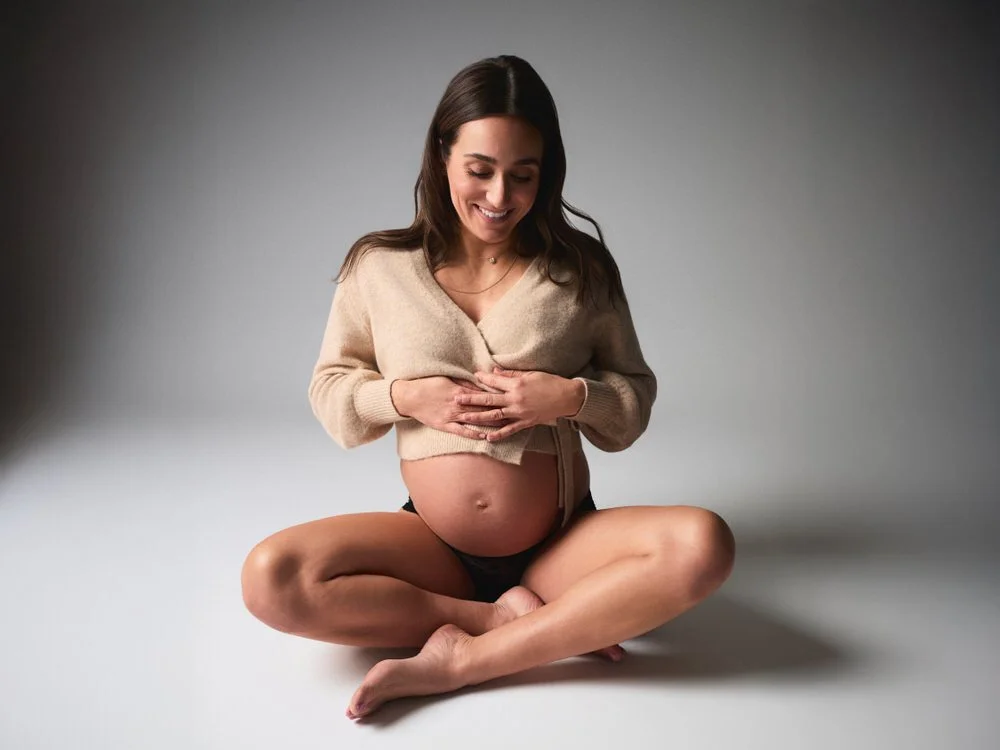 Expectant mother seated cross-legged in a beige sweater, smiling down at her belly in a softly lit maternity portrait.