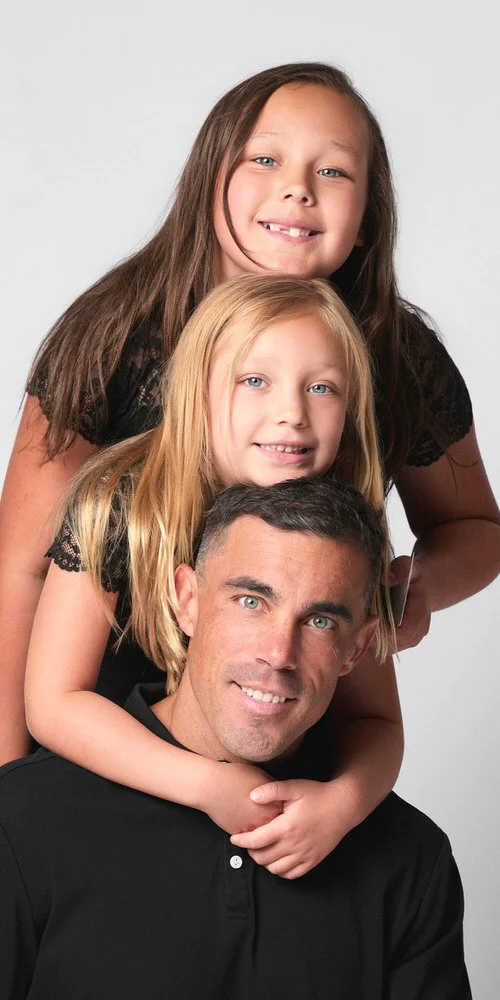 Studio family portrait — dad with two daughters stacked in a vertical pose, all in black