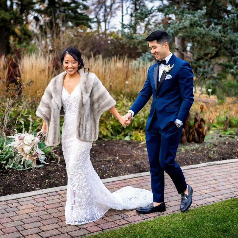 Outdoor wedding portrait — couple walking through a garden, bride in a fur coat, groom in a blue suit