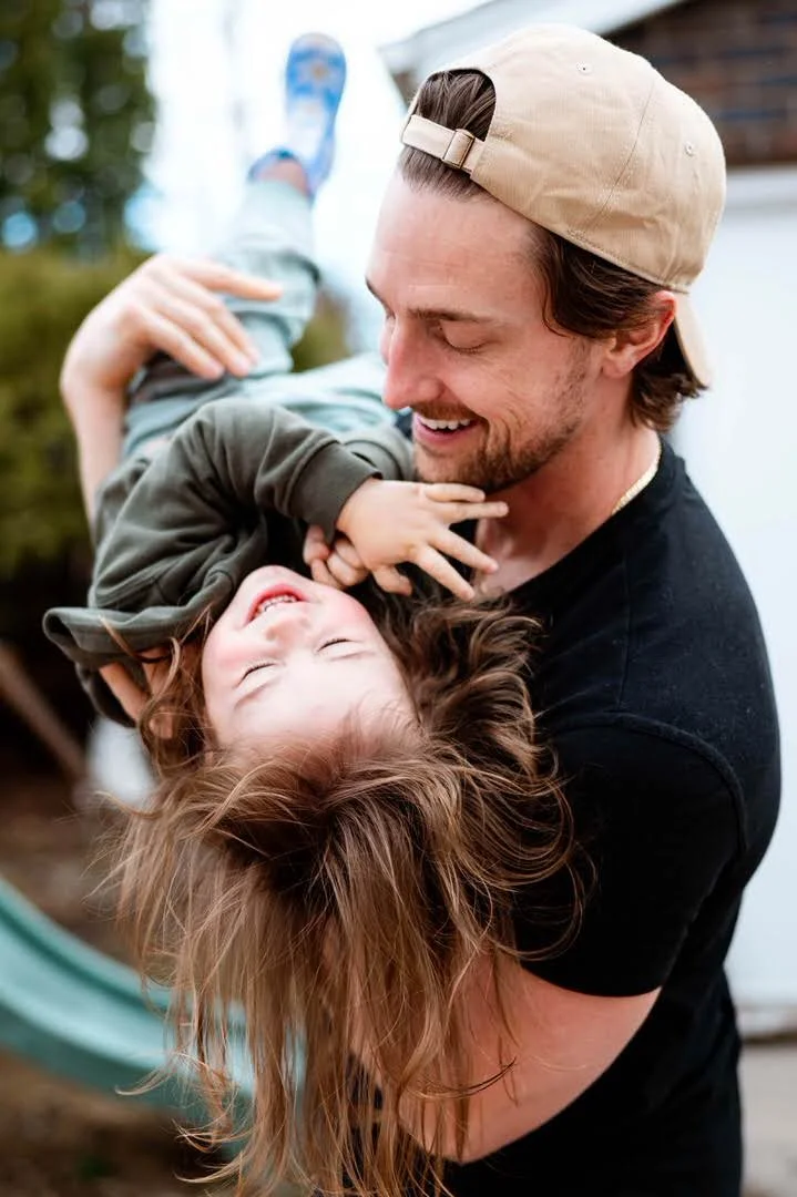 Mark Eadie holding his laughing toddler daughter upside down outdoors