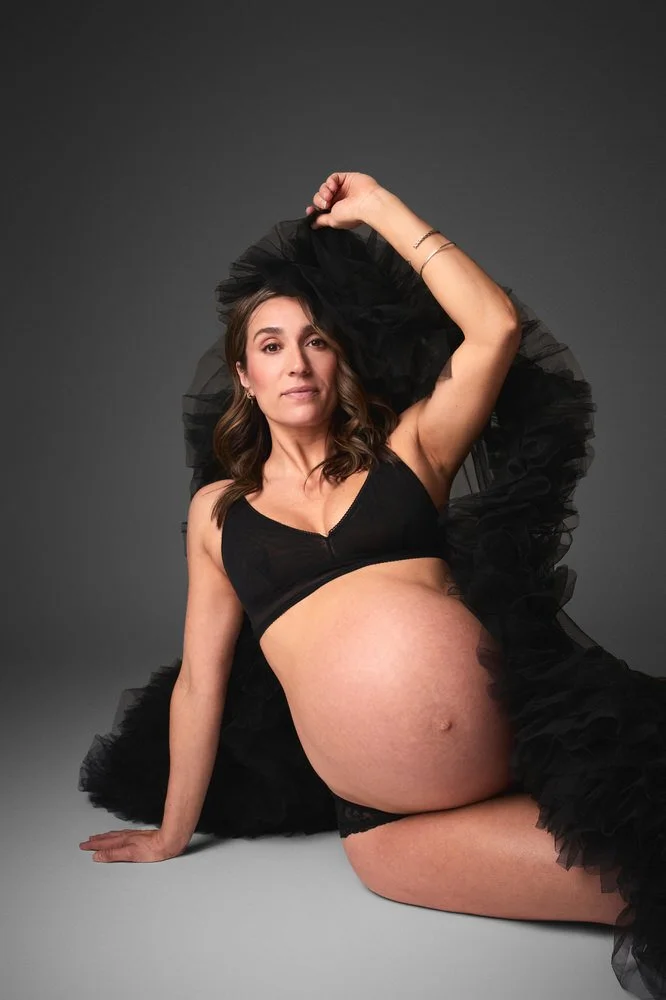 Expectant mother seated on the studio floor in a black tulle robe, one arm lifted
