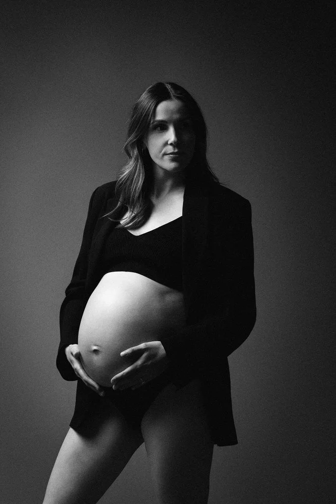Black-and-white maternity portrait, an expectant mother in an open black blazer cradling her belly in dramatic, low studio light.