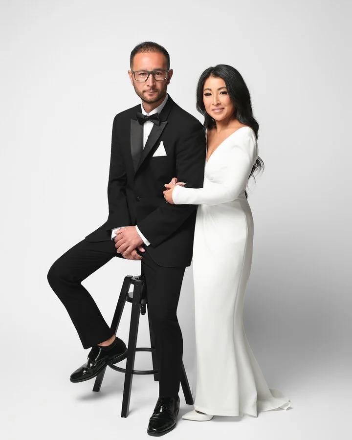 Studio wedding portrait — couple full-length on stool against a white backdrop