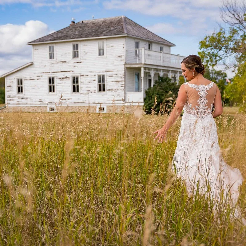 Outdoor bridal portrait — bride standing in a field with an old house in the background