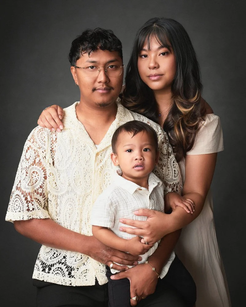 Studio family portrait of two parents and their toddler son, dressed in cream and lace, photographed against a dark backdrop at our Exchange District studio.