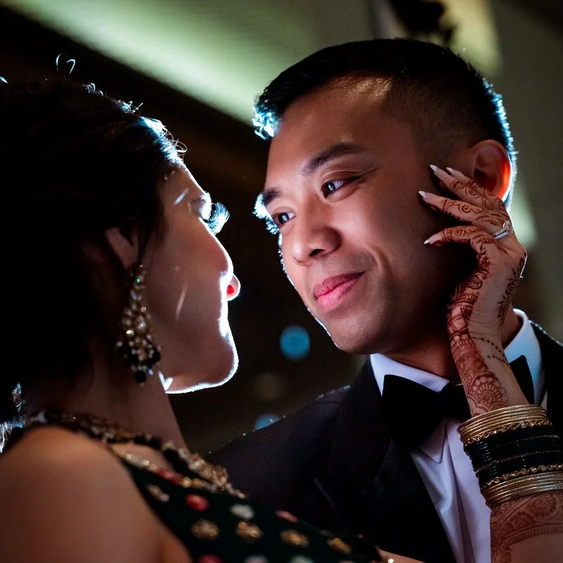 Close-up evening portrait of a bride resting her henna-painted hand on her groom's cheek, string lights glowing softly behind them.