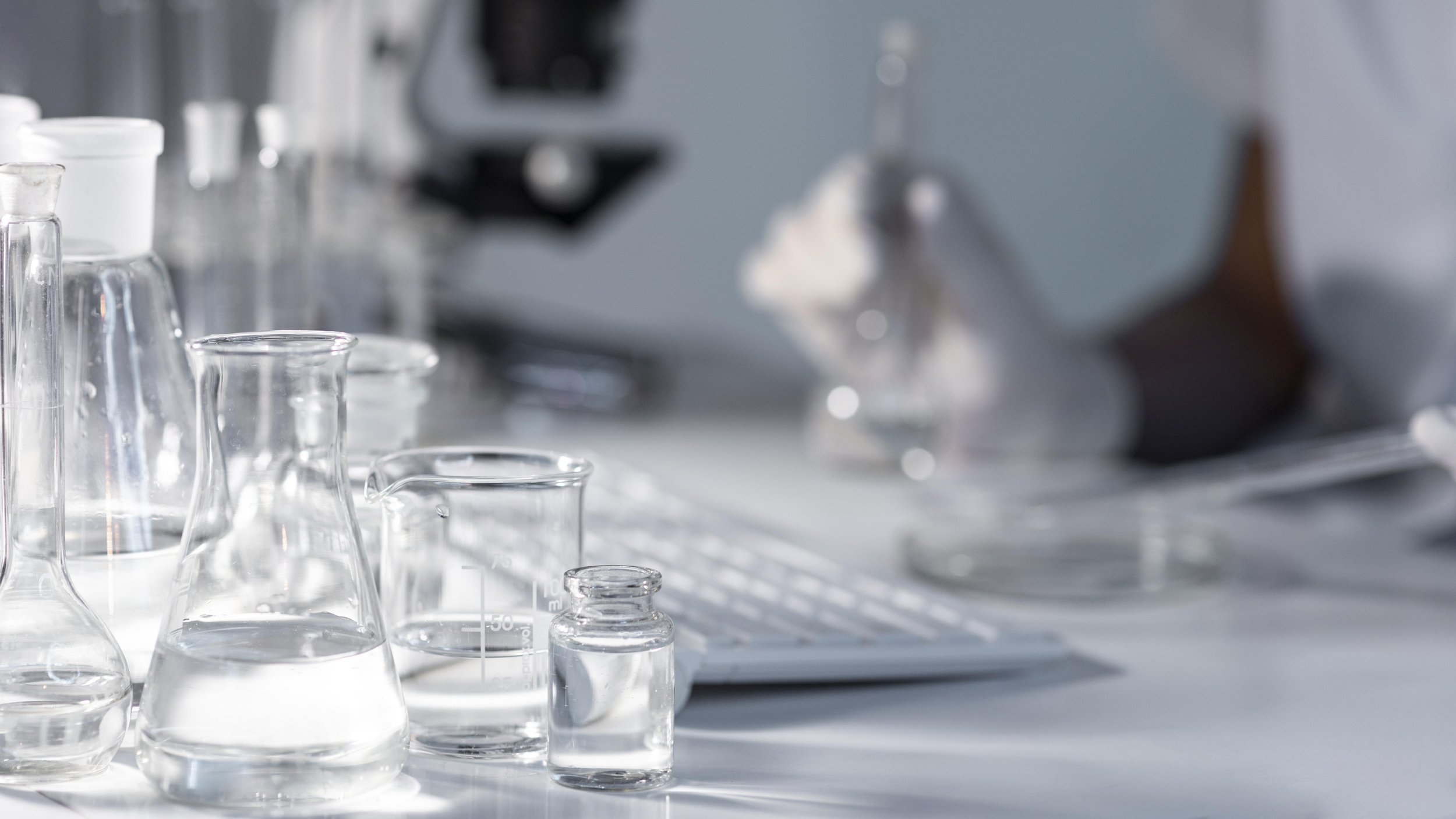 closeup of beakers filled with clear solution on a desk with gloved hands pipetting solution into petri dish in the background