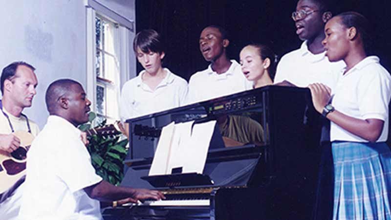 Photo of students gathered around upright piano singing.